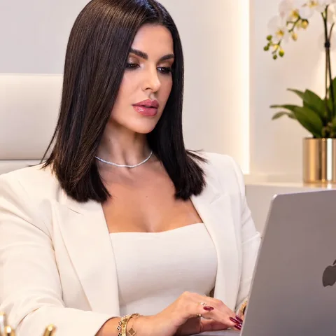 Dra. Solyanni Abreu à mesa do consultório, de blazer branco, atendendo pelo notebook